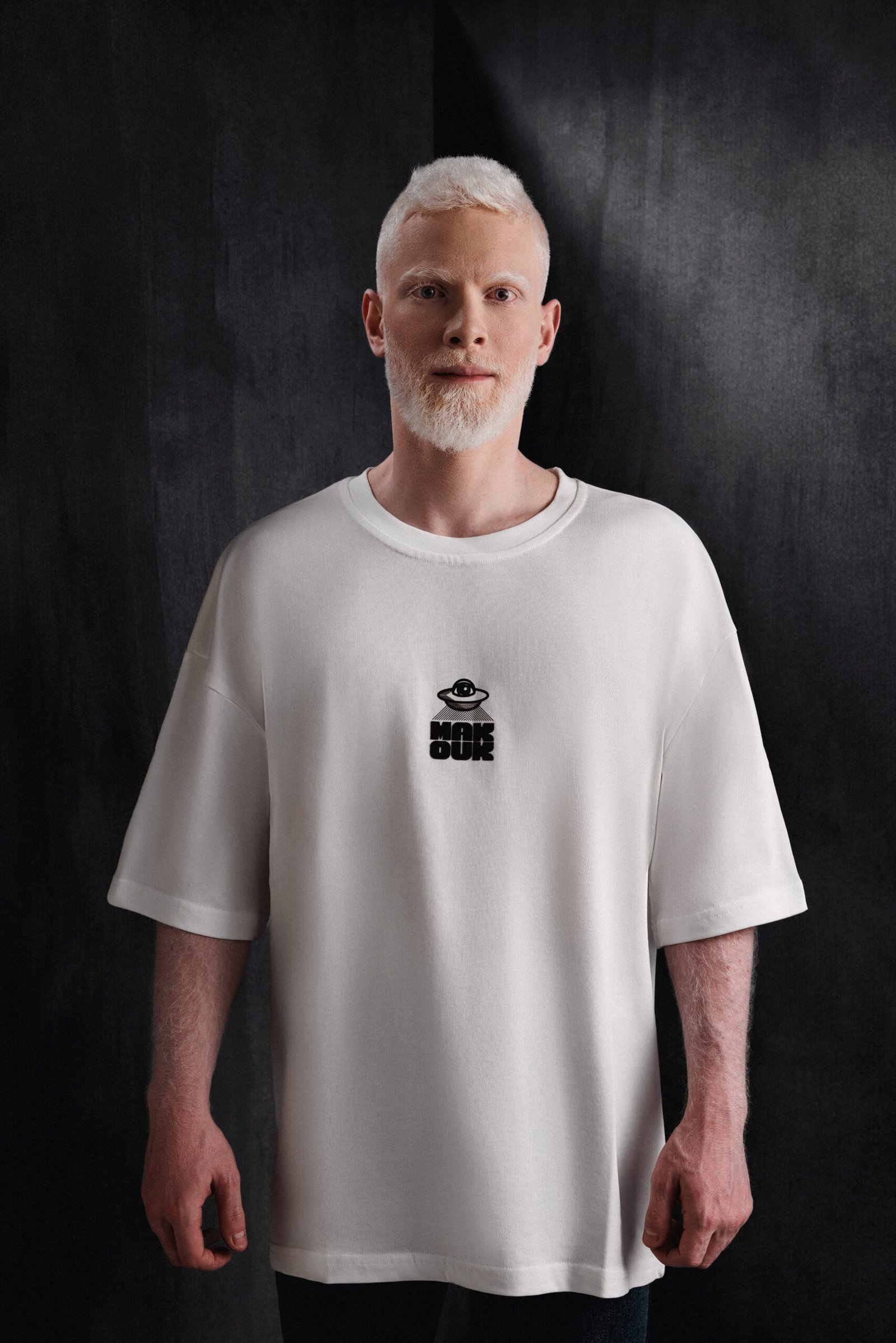 Model in oversized white Makouk t-shirt with black UFO logo, standing against dark backdrop. Designed by Mohamad Ali Makouk for Makouk Studios.
