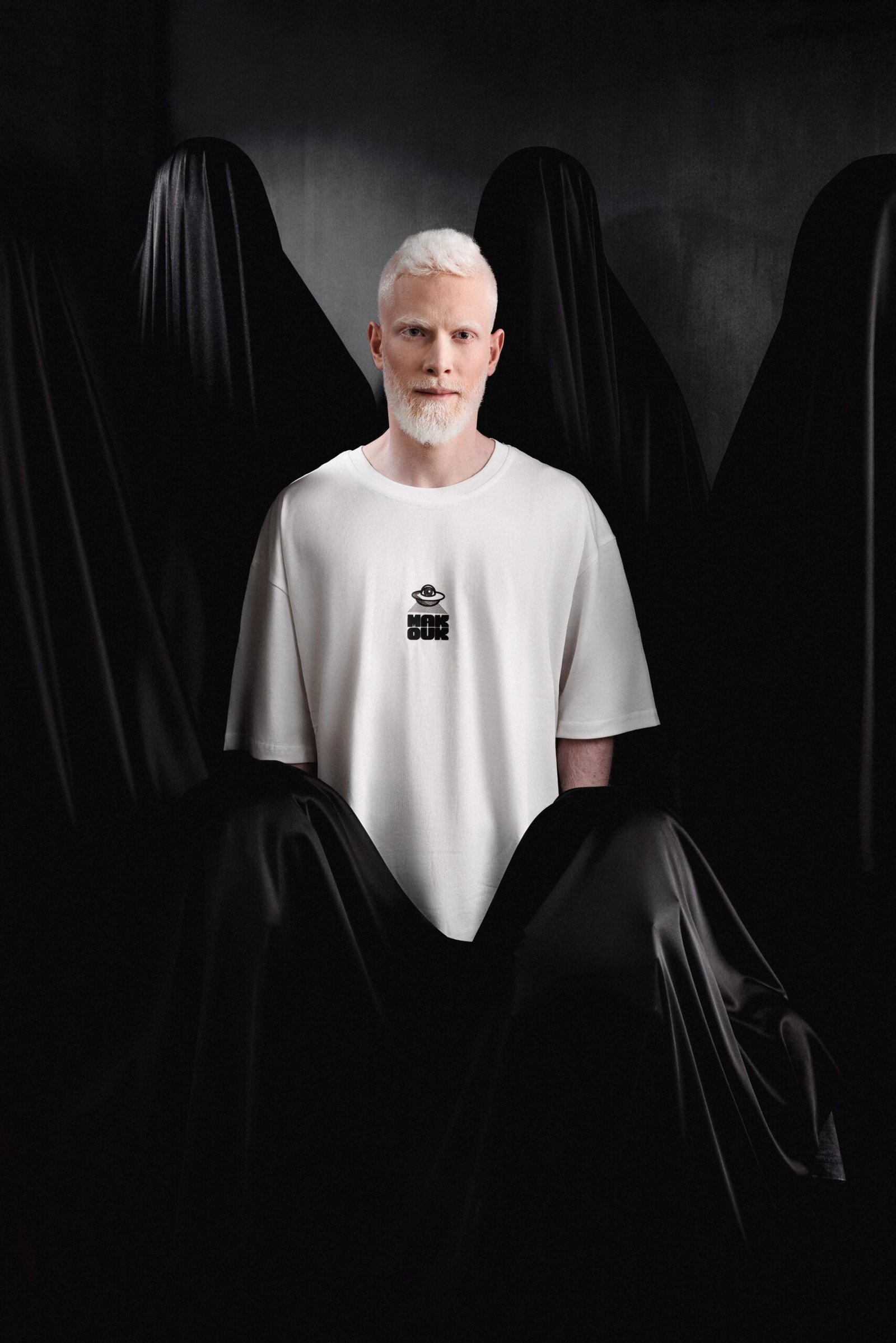 White male model with platinum blonde hair wearing a white oversized Makouk Studios t-shirt with black logo, surrounded by figures draped in black veils. From Macho Meltdown Collection by Mohamad Ali Makouk.