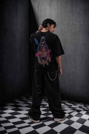 Back view of Makouk Studios black oversized graphic T-shirt featuring a surreal purple hooded figure carrying a pink child, styled with baggy black jeans in a contemporary streetwear editorial.